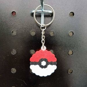 May include: A red, black, and white beaded keychain in the shape of a Poké Ball. The keychain is attached to a silver metal ring and chain. The background is a black pegboard.
