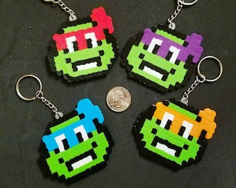 Pode incluir: Quatro chaveiros de arte de pixel com as Tartarugas Ninja. Cada chaveiro é de uma cor diferente: vermelho, roxo, azul e laranja. Os chaveiros são feitos de contas de plástico e têm um contorno preto.