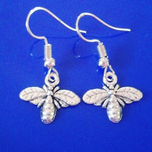 May include: A pair of silver-colored bee earrings. Each earring features a detailed bee charm with outstretched wings, suspended from a simple hook. The earrings are set against a vibrant blue background.
