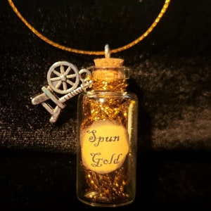 May include: A gold necklace with a small glass bottle charm filled with gold thread and a spinning wheel charm. The bottle has a label that reads "Spun Gold".