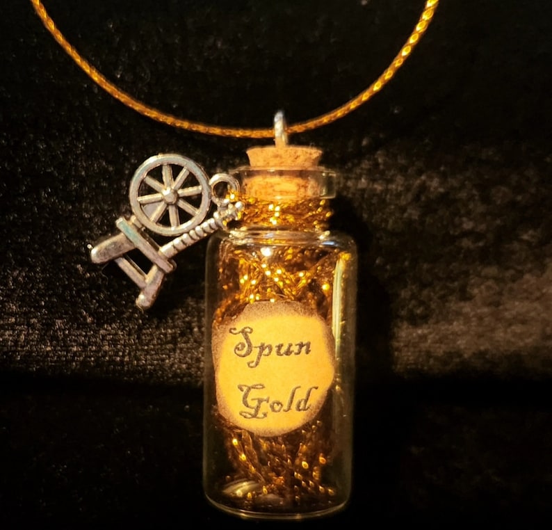 May include: A gold necklace with a small glass bottle charm filled with gold thread and a spinning wheel charm. The bottle has a label that reads "Spun Gold".