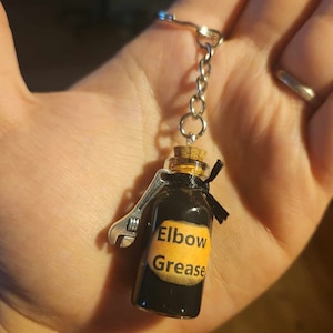May include: A black glass bottle with a cork stopper and a silver wrench charm. The bottle has a label that says "Elbow Grease".