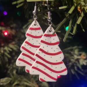May include: A pair of white Christmas tree earrings with red and green stripes and sprinkles. The earrings are made of polymer clay and have silver hooks.
