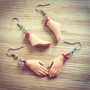 Op de afbeelding: Drie paar oorbellen met een afgehakte hand en voet. De oorbellen zijn gemaakt van polymer clay en hebben een realistische uitstraling. De hand en voet zijn rood geverfd op de plaats van de snee.