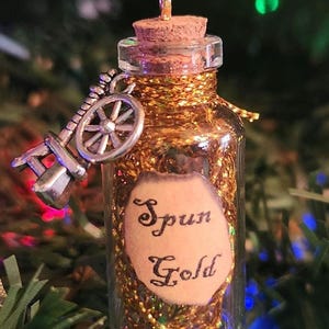 May include: A small glass bottle filled with gold glitter and a cork stopper. The bottle is hanging from a gold chain and has a silver spinning wheel charm. A label on the bottle reads "Spun Gold".