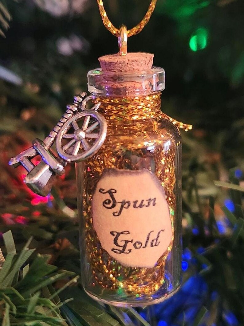 May include: A small glass bottle filled with gold glitter and a cork stopper. The bottle is hanging from a gold chain and has a silver spinning wheel charm. A label on the bottle reads "Spun Gold".