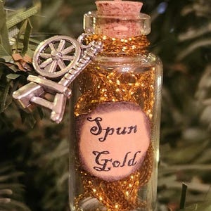 May include: A small glass bottle with a cork stopper filled with gold glitter and a spinning wheel charm. The bottle has a label that reads "Spun Gold".