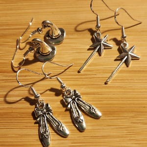 May include: A set of six silver earrings with different charms. The charms include a witch's hat, a magic wand with a star, and a pair of ballet shoes.