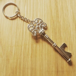 May include: Silver-colored key-shaped keychain with an ornate design. The key has a decorative top and a bottle opener at the end. The keychain is attached to a metal ring and chain. The background is a light brown wooden surface.