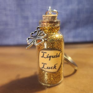 May include: A small glass bottle filled with gold glitter and a cork stopper. The bottle has a silver charm attached to it in the shape of a four-leaf clover. The bottle has a label that reads "Liquid Luck".
