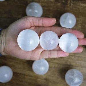 May include: A hand cradles three polished, white selenite spheres, highlighting their smooth, translucent appearance. Other spheres are on a wooden surface. These spheres are a natural, decorative item.