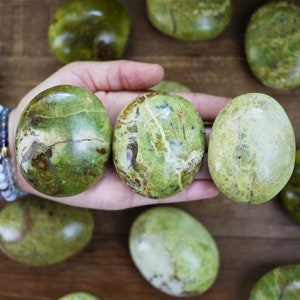 Peut inclure: Trois pierres d'opale verte polies sont tenues dans une main, avec plusieurs autres dispersées sur une surface en bois. Les pierres ovales présentent des nuances de vert et des veines brunes. Des bracelets sont visibles en arrière-plan.