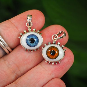 Puede incluir: Dos colgantes con forma de ojo con engastes de metal plateado. Uno tiene un iris azul, el otro un iris naranja, ambos con pupilas negras. Los colgantes están sobre un fondo blanco y tienen un pequeño lazo en la parte superior para una cadena.