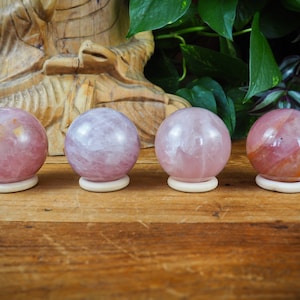 Può includere: Quattro sfere di quarzo rosa, ciascuna su un piccolo supporto bianco sporco. Le sfere variano nelle tonalità di rosa e sono esposte su una superficie di legno. Lo sfondo include una figura di legno intagliata e fogliame verde.