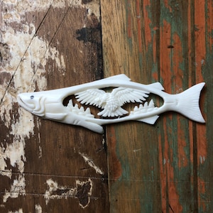 Puede incluir: Escultura de pez tallada en blanco roto con un águila y una escena de montaña recortada en el centro. El pez está sobre una superficie de madera desgastada y multicolor. El águila tiene las alas extendidas.