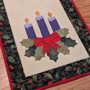 Peut inclure: Chemin de table rectangulaire de Noël avec un centre crème orné d'un motif de houx et de bougies. Les bougies sont violettes avec des flammes orange, le houx a un nœud rouge. La bordure est noire avec du houx et des baies rouges.