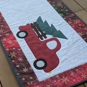 May include: A festive Christmas table runner with a red truck design carrying a green Christmas tree. The runner has a white central panel with the truck and tree applique, framed by a red border with snowflake patterns. The runner is approximately 152 cm long.