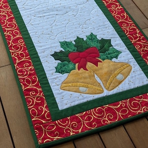 Peut inclure: Chemin de table rectangulaire de Noël. Le centre est bleu clair avec un motif matelassé. Des cloches dorées, des nœuds rouges et des feuilles de houx vertes sont appliqués aux extrémités. La bordure est rouge avec des volutes dorées et une bordure verte.