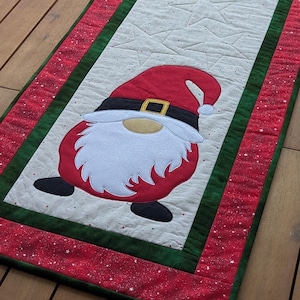 Könnte beinhalten: Ein rechteckiger Weihnachtstischläufer mit zwei Zwergenmotiven. Die Zwerge haben rote Hüte, weiße Bärte und schwarze Schuhe. Der Läufer hat eine weiße Mitte mit Sternsteppung, umrandet von grünen und roten Stoffen.