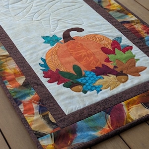 May include: A quilted table runner with a cream-colored center featuring leaf patterns. The runner has a brown border and is decorated with appliqued pumpkins and colorful leaves, acorns, and berries. The runner is placed on a wooden table.