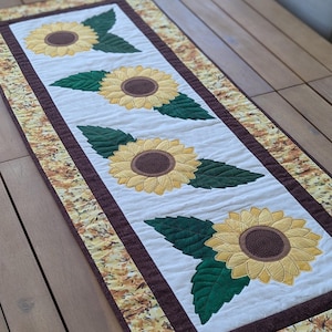 Peut inclure: Chemin de table rectangulaire avec quatre tournesols brodés et des feuilles vertes sur fond blanc. Bordure marron et bordure à motifs de feuilles jaunes et marron. Posé sur une table en bois.
