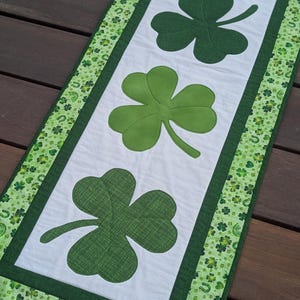 May include: A St. Patrick's Day table runner featuring four green shamrocks on a white background. The runner is bordered with a green fabric patterned with shamrocks and horseshoes. The runner is approximately 12 inches wide and 36 inches long.