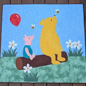 May include: A handmade quilt featuring Winnie the Pooh and Piglet. Pooh is yellow, Piglet is pink, and they are sitting on a log. A red balloon floats in the sky, with bees and white flowers in the background. The quilt is square.