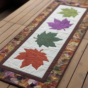 Könnte beinhalten: Rechteckiger Tischläufer mit applizierten Ahornblättern in Orange, Grün und Lila auf cremefarbenem Hintergrund. Der Läufer hat einen braunen Rand und einen gemusterten Blattrand. Auf einem Holztisch platziert.