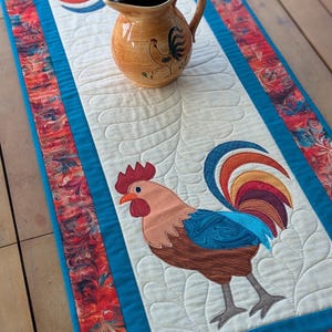 May include: A rectangular quilted table runner featuring two rooster appliqués with brown, blue, and red plumage, set against a cream background with feather-like stitching. A tan ceramic pitcher with a painted rooster motif sits on top.