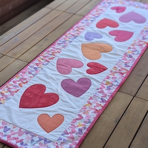 May include: A rectangular table runner featuring a white center with appliqued hearts in shades of red, pink, and orange. The border has a watercolor heart pattern with a pink trim. The runner is placed on a wooden surface.