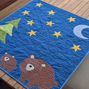 Puede incluir: Una manta acolchada azul con un diseño de cielo nocturno, con estrellas amarillas, una luna creciente blanca y un árbol verde. Dos osos marrones están aplicados en la parte inferior. La manta está sobre una mesa de madera.