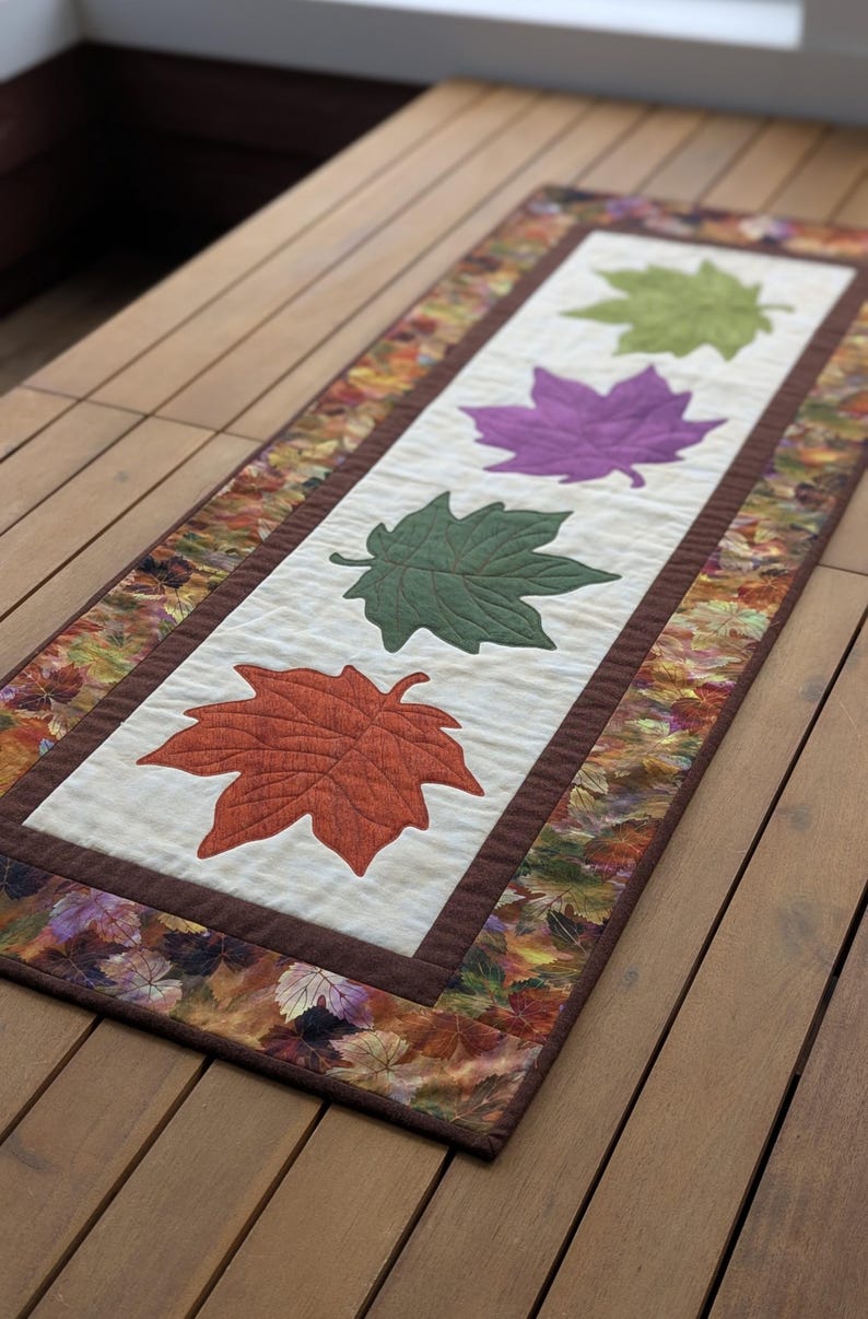 May include: A rectangular table runner featuring a central panel with four appliqued maple leaves in orange, green, and purple. The leaves are set against a cream background, framed by a brown border and a patterned leaf-print edge.