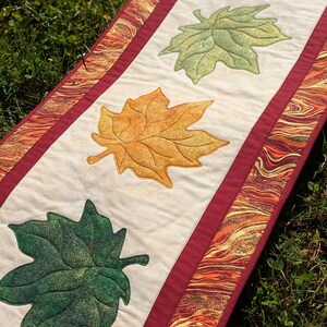 May include: A rectangular fall-themed table runner with four embroidered maple leaves in shades of green, yellow, and orange. The leaves are centered on a cream-colored fabric, bordered by a maroon band and a patterned edge.