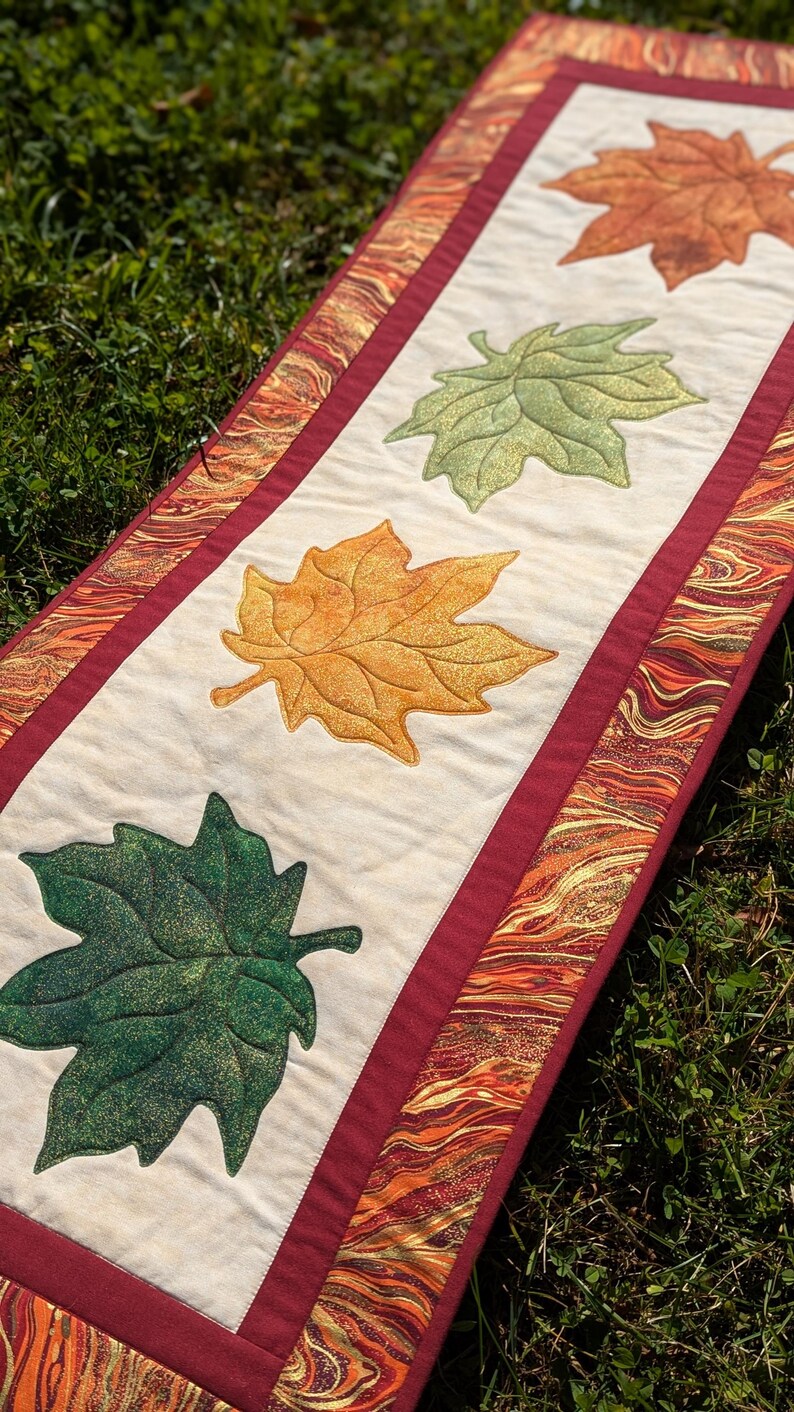 May include: A rectangular fall-themed table runner with four embroidered maple leaves in shades of green, yellow, and orange. The leaves are centered on a cream-colored fabric, bordered by a maroon band and a patterned edge.