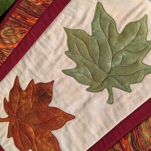 May include: A quilted table runner featuring two large appliqued leaves, one green and one brown, on a cream-colored background. The runner is bordered with a burgundy fabric and a patterned fabric with orange, gold, and brown swirls.