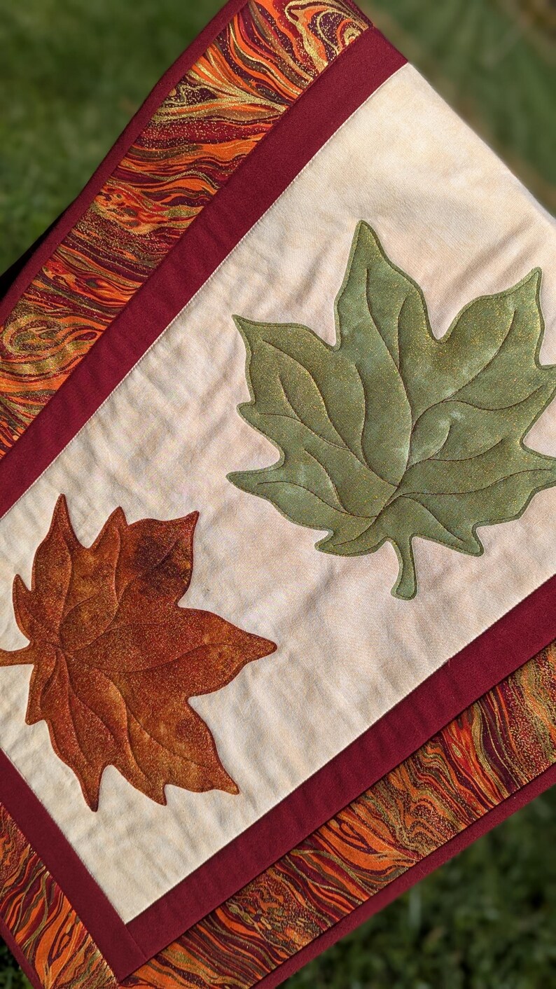 May include: A quilted table runner featuring two large appliqued leaves, one green and one brown, on a cream-colored background. The runner is bordered with a burgundy fabric and a patterned fabric with orange, gold, and brown swirls.