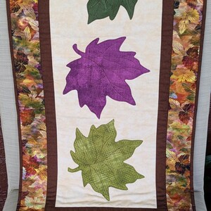 May include: A fall-themed table runner featuring three large leaf appliqu&eacute;s in green and purple on a cream background. The edges are bordered with a brown fabric and a leaf pattern.