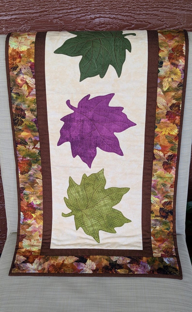 May include: A fall-themed table runner featuring three large leaf appliqu&eacute;s in green and purple on a cream background. The edges are bordered with a brown fabric and a leaf pattern.