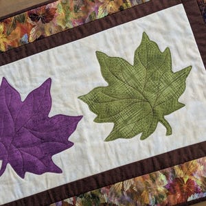 May include: A quilted table runner featuring three large leaf appliqu&eacute;s in green and purple on a cream background. The runner is bordered with a brown fabric and a fall leaf pattern, perfect for autumn decor.