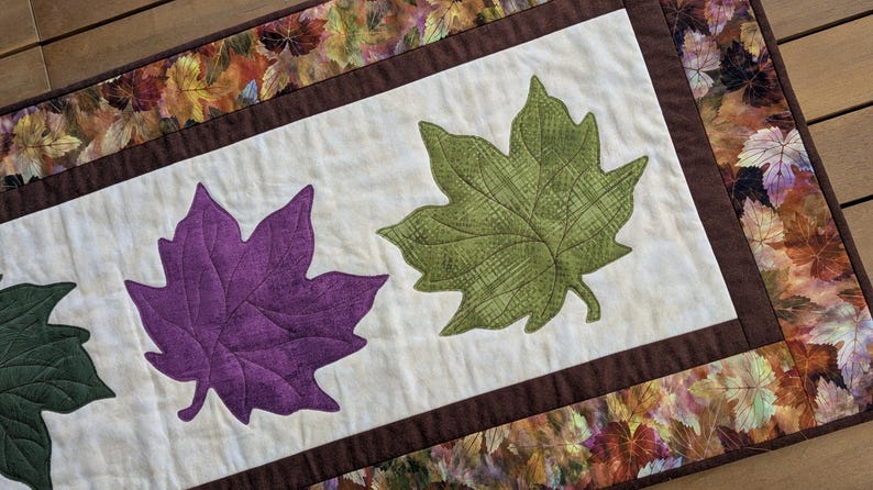 May include: A quilted table runner featuring three large leaf appliqu&eacute;s in green and purple on a cream background. The runner is bordered with a brown fabric and a fall leaf pattern, perfect for autumn decor.