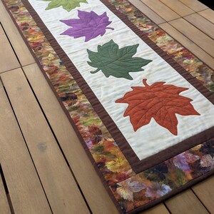 May include: A rectangular table runner featuring four appliqued maple leaves in green, purple, green, and orange. The leaves are centered on a cream-colored background, framed by a brown border and a fall-themed leaf print.