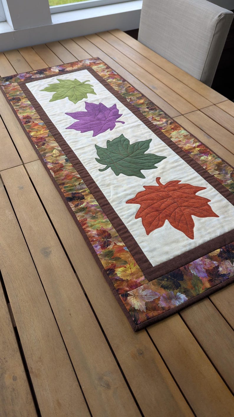 May include: A rectangular table runner featuring four appliqued maple leaves in green, purple, green, and orange. The leaves are centered on a cream-colored background, framed by a brown border and a fall-themed leaf print.