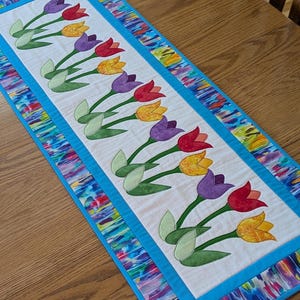 Puede incluir: Camino de mesa rectangular con diseño floral. El centro del camino de mesa es blanco con tulipanes aplicados en rojo, amarillo y morado. El borde es una vibrante mezcla de colores, enmarcado por un borde azul brillante. Aproximadamente 152 cm de largo.