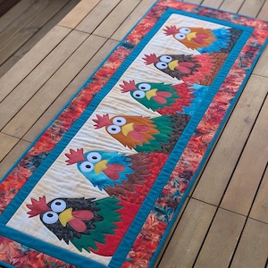 May include: A rectangular fabric table runner featuring a cream-colored center panel with six appliqued roosters in various colors including blue, green, brown, and black. The border is a vibrant red and orange marbled pattern with teal trim.