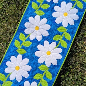 May include: A rectangular table runner featuring a mottled blue background with a repeating pattern of white daisy flowers with yellow centres and bright green stems and leaves, finished with a vibrant lime green border trim along the edges.