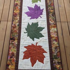 May include: A rectangular table runner featuring four large, colorful leaf appliqu&eacute;s in green, purple, dark green, and orange-red. The leaves are centered on a cream-colored background, framed by a brown border and a fall-themed leaf print.