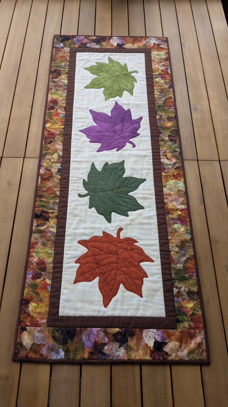 May include: A rectangular table runner featuring four large, colorful leaf appliqu&eacute;s in green, purple, dark green, and orange-red. The leaves are centered on a cream-colored background, framed by a brown border and a fall-themed leaf print.