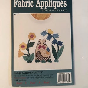 May include: A white t-shirt with an iron-on appliqué kit featuring a cat in a garden with flowers. The kit includes die-cut appliqué shapes with Fus-O-Bond already applied to complete one project. The text on the packaging reads "Fabric Appliqués Iron-On Appliqué Kit 55130 Garden Kitty Past! Fun! Easy! Made in U.S.A."
