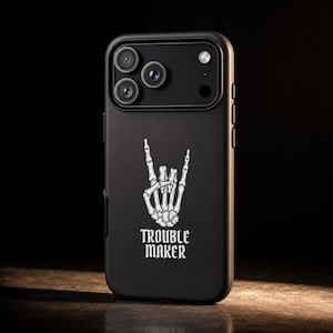 May include: A matte black smartphone case featuring a white graphic of a skeleton hand making a rock and roll sign. Below the hand, the text TROUBLE MAKER is printed in a bold, stylized font. The case includes a cutout for a triple camera system.