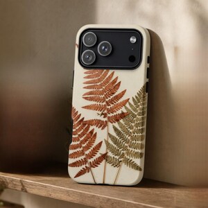 Può includere: Una cover per smartphone color crema con una stampa botanica di foglie di felce pressate nei toni dell'arancione ruggine e del verde oliva spento. La cover presenta un ritaglio rettangolare nero per la fotocamera con tre lenti e un flash.